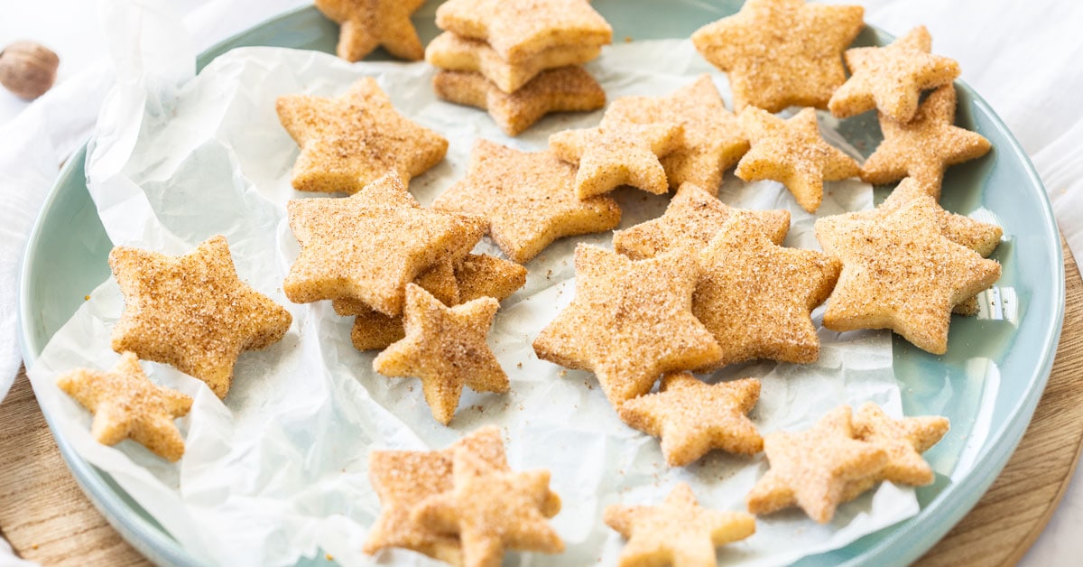 Un plato verde pastel cubierto con galletas de mantequilla en forma de estrella. Una taza de t&eacute; al fondo
