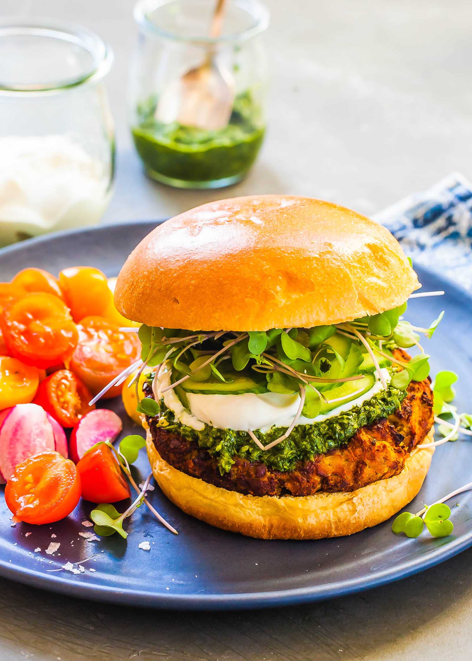 Burger de lentilles avec chutney repose sur une assiette bleue avec un côté de tomates raisins coupées en deux. Un petit plat de chaque yaourt et chutney est visible au-dessus du burger.