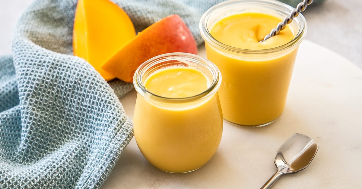 Un peque&ntilde;o frasco de conserva lleno de cuajada de mango amarillo, con otro frasco y rebanadas de mango detr&aacute;s.