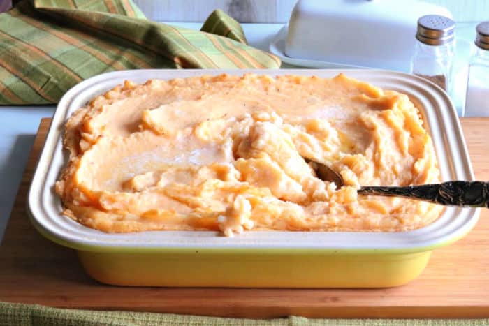 A horizontal photo of a yellow casserole dish of cheesy horseradish mashed potatoes casserole with a spoon.