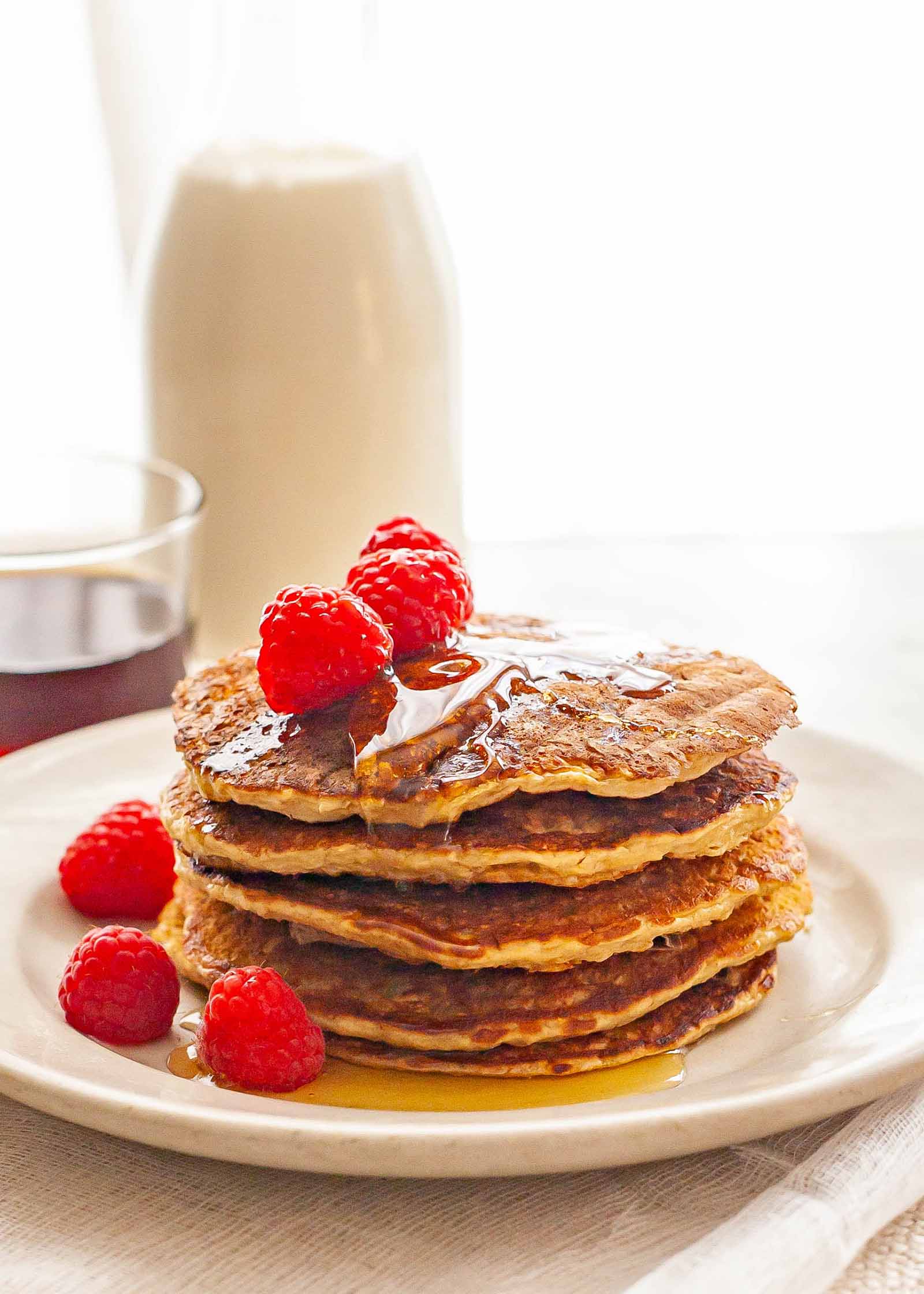 Crêpes à l'avoine sans gluten empilées haut sur une assiette de whie arrosée de sirop d'érable et garnie de framboises et d'une bouteille en verre de lait derrière la pile.