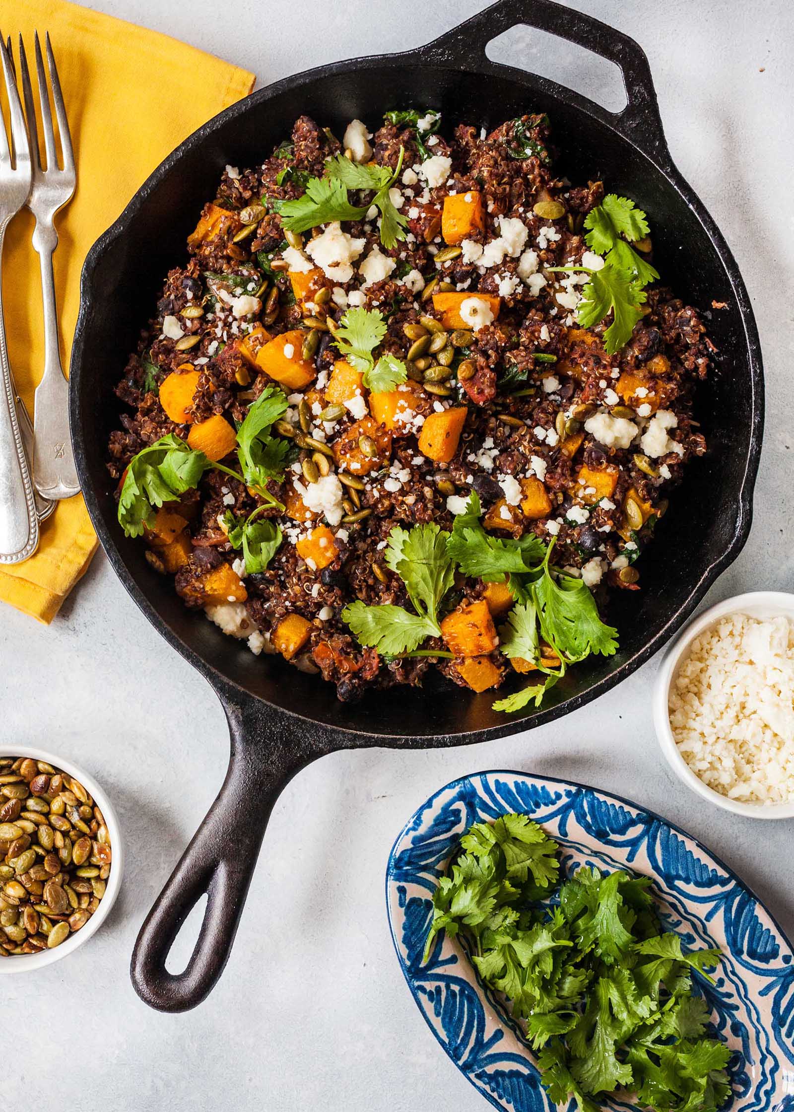 Vue de dessus d'une poêle en fonte de dîner végétarien avec des graines de citrouille, du quinoa, de la courge musquée et de la coriandre visible dans le pot. Dans le sens des aiguilles d'une montre autour du pot se trouve un petit bol de riz blanc, un petit plateau de coriandre, un ramiken de graines de citrouille grillées et une serviette jaune avec deux fourchettes côte à côte.