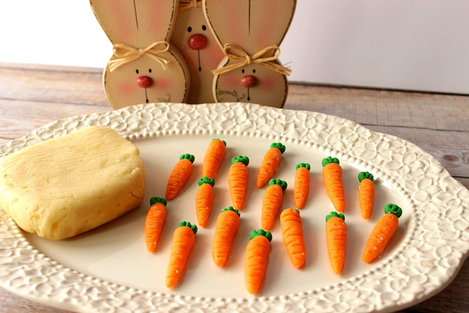 A white platter with a block of homemade marzipan and marzipan carrots, with bunnies in the background.