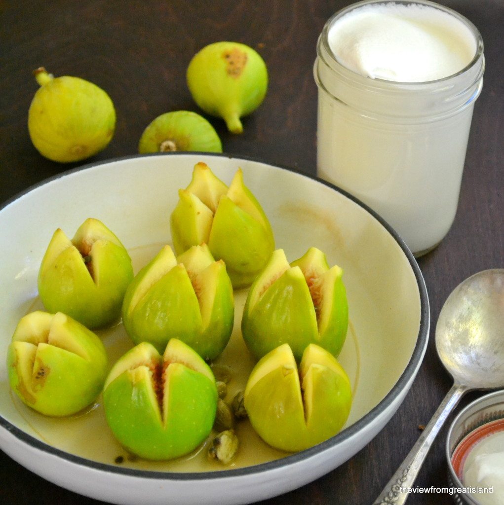 Foto de higos para cardamomo y brandy higos escalfados en una sartén blanca, con un tarro de crema batida y algunos higos más en el fondo.
