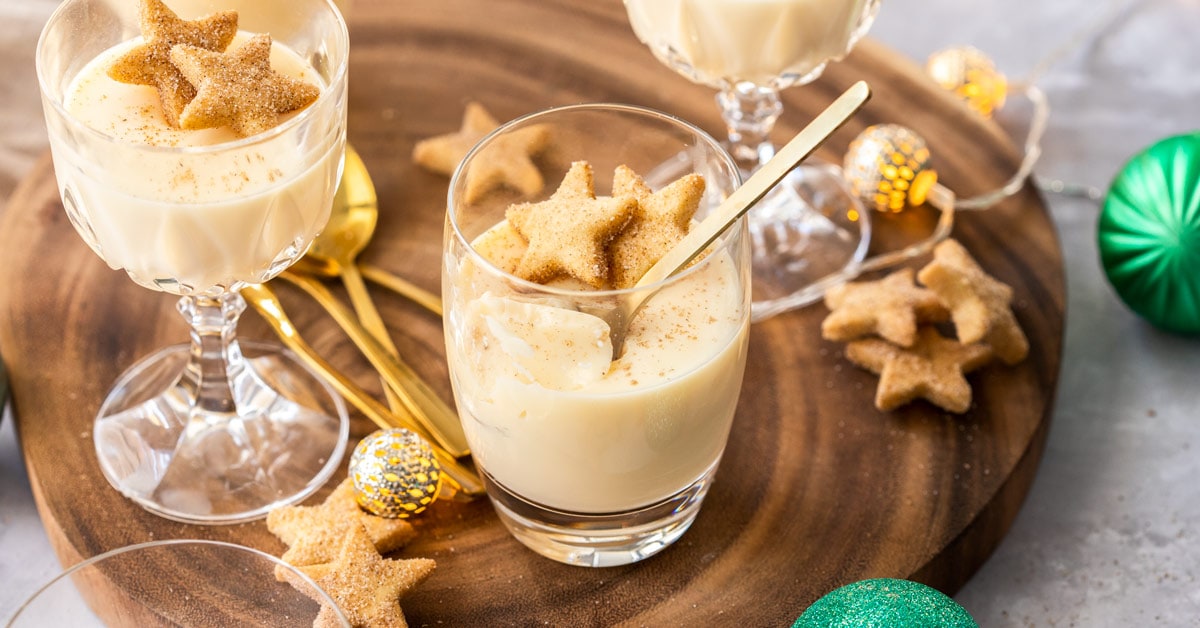 Vasos rellenos de ponche de huevo panna cotta sobre una tabla de madera, rodeados de galletas de mantequilla y adornos festivos.