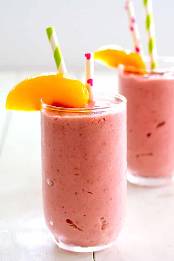Dos vasos transparentes rellenos de batido de frambuesa, melocot&oacute;n y reques&oacute;n rosa adornado con pajitas y una rodaja de durazno