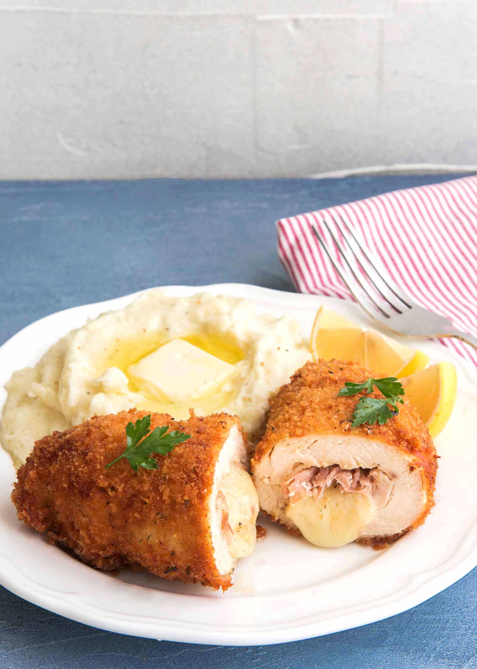 Best Chicken Cordon Bleu on a white plate with mashed potatoes and butter, a silver fork and a red and white stripped napkin against a blue background.
