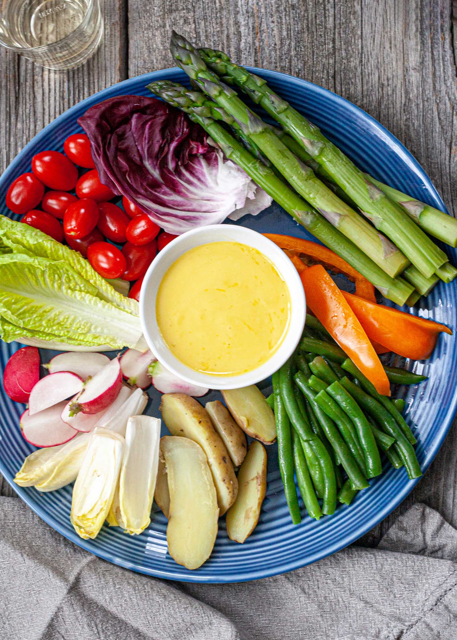 A plate with homemade aioli in a small bowl in the center of a plate. The aioli sauce is surrounded by a variety of steamed and fresh vegetables.
