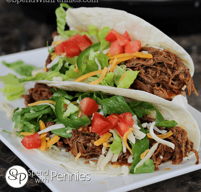 two soft shelled tacos filled with shredded beef and vegetables with a slow cooker in the background