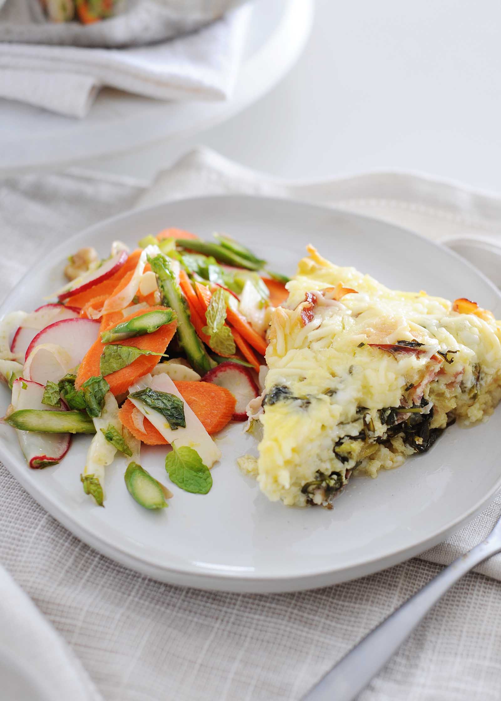 Side view of a slice of breakfast casserole on a white plate with a spring salad with sliced carrots, radishes, asparagus and mint mixted together. Greens are visible in the slice of strata. Cream linens are under the plate and a fork is partially visible in the lower right corner. The skillet and linens are partially in view in the upper left corner.