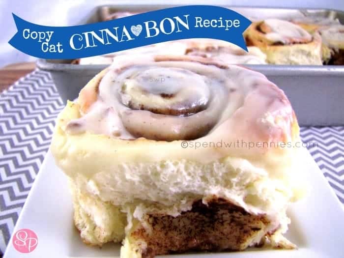 single copy cat cinnabon with frosting on a white plate with a tray of cinnamon buns in the background