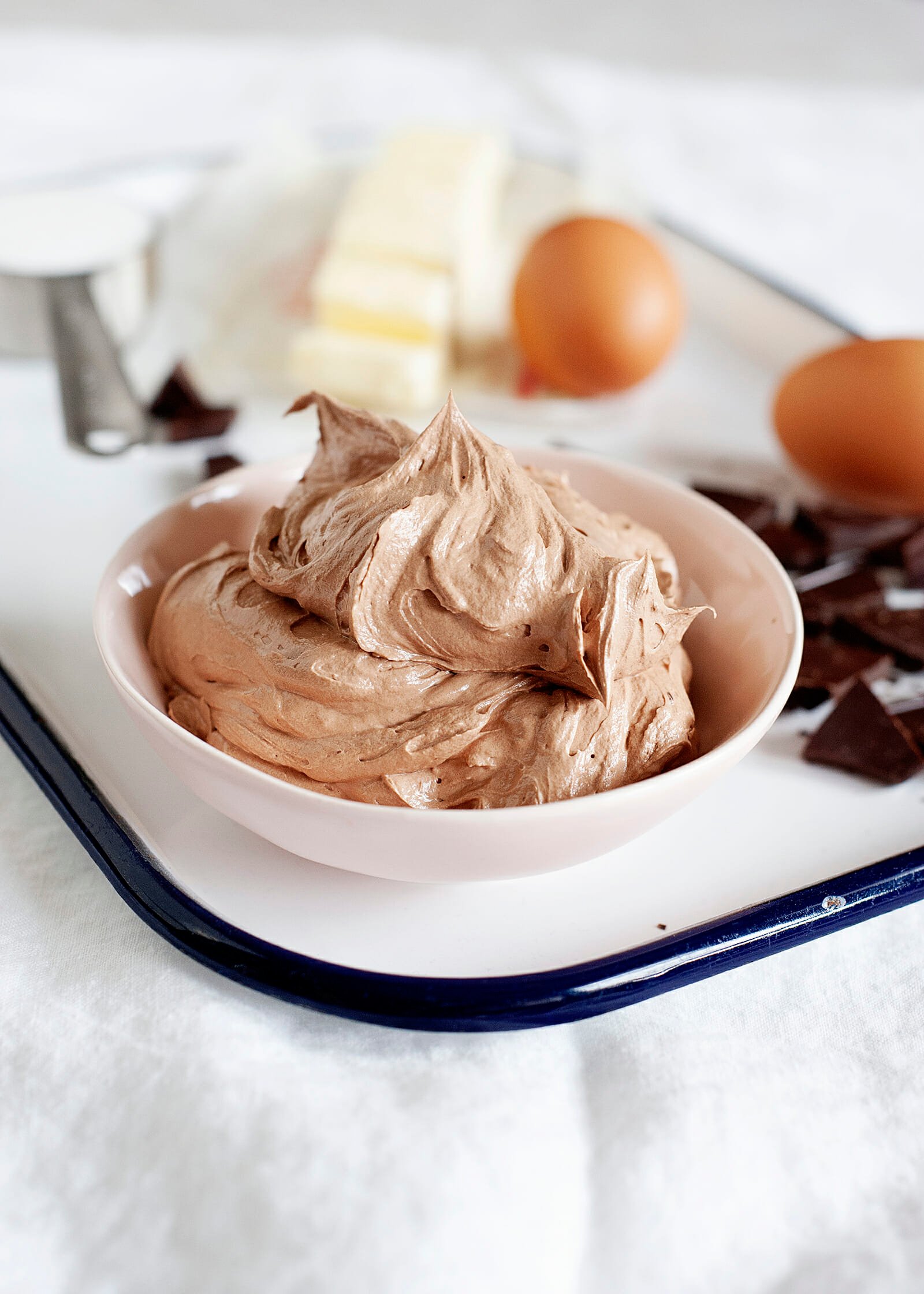 Perfect Chocolate Swiss Meringue Buttercream Piled into a pink bowl and set on a white tray with butter, chocolate pieces and sugar around it.