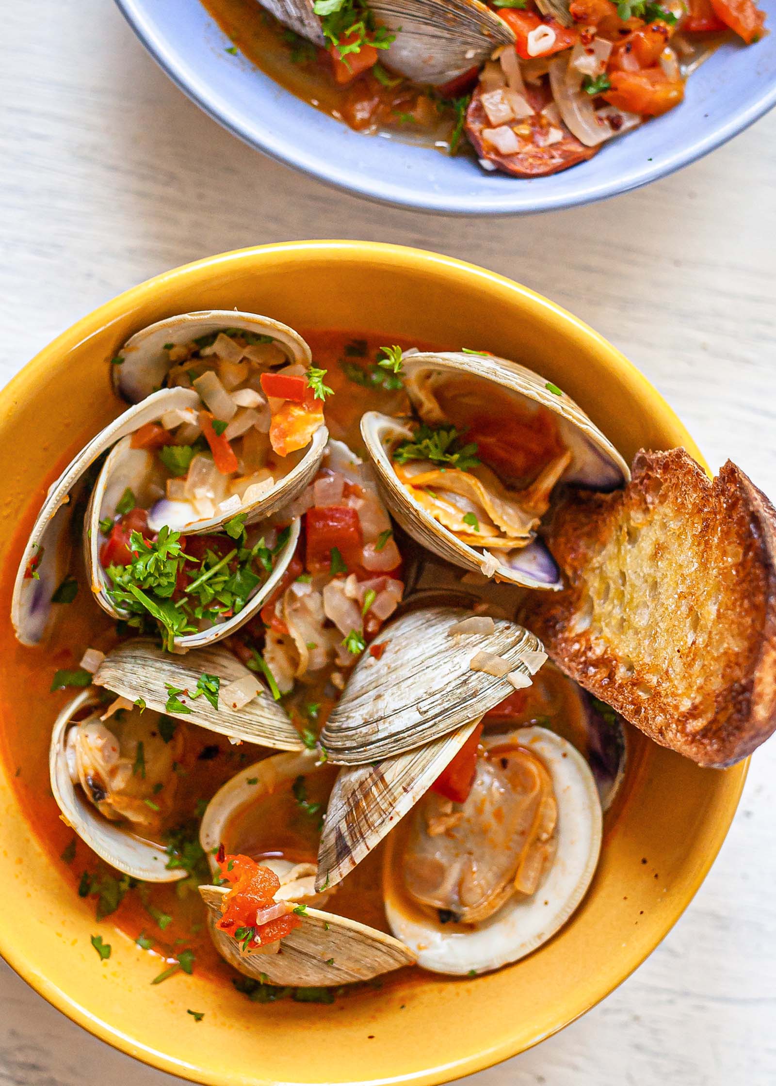 Overhead view of a yellow bowl with steamed clams with chorizo and grilled bread. A second blue bowl is partially visible above.