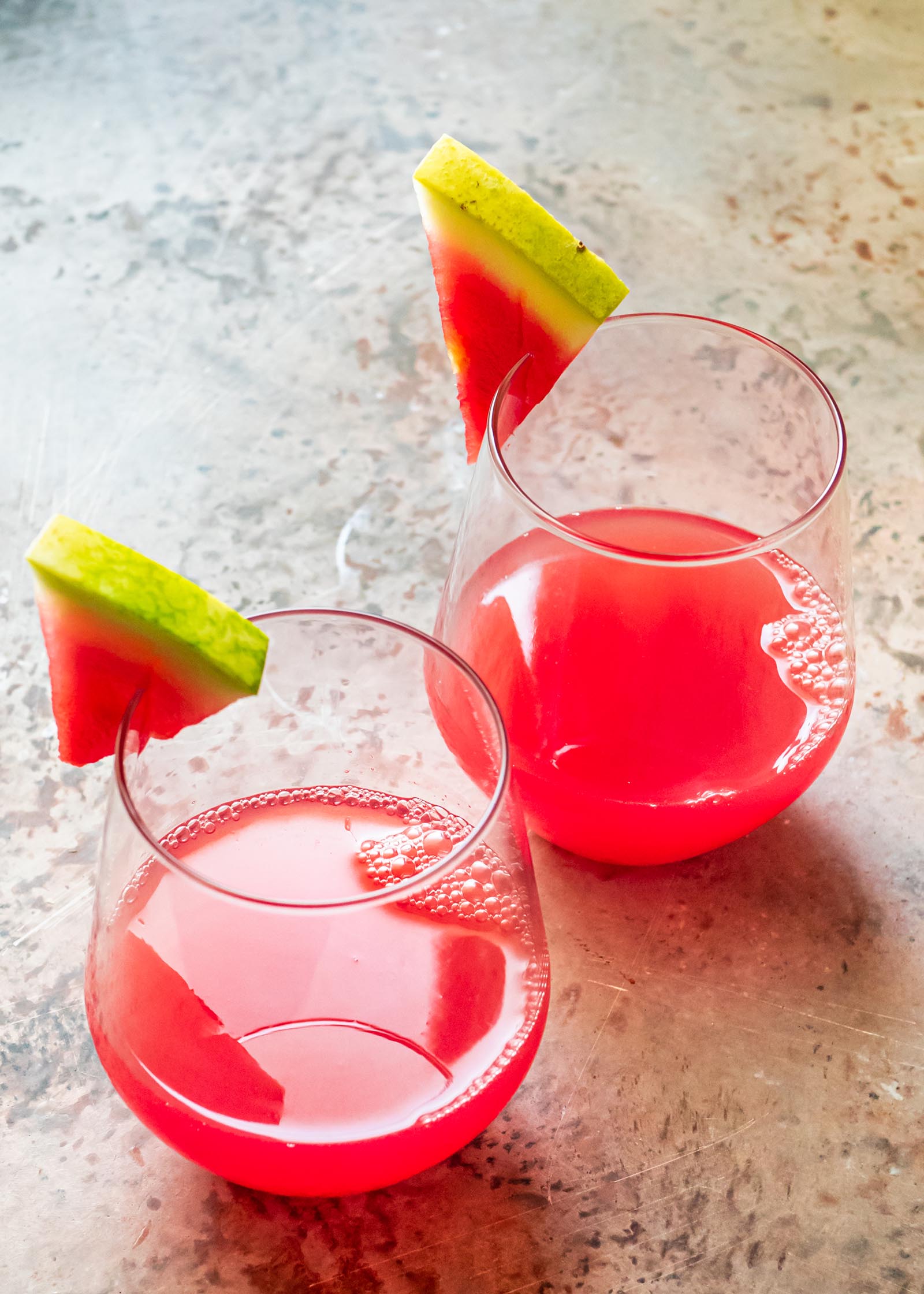Two wine glasses with watermelon drinks inside and a thin slice of watermelon on the edge.
