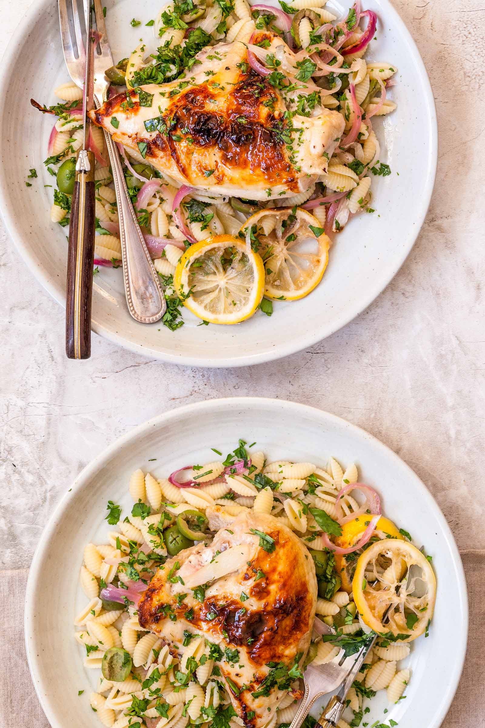 A vertical photo of two plates with easy weeknight chicken on each. Each plate has a golden brown roasted chicken breast, roasted lemon wedges, sliced red onion, shell pasta and herbs scattered over top. The top plate has a knife and fork on the left side of the plate. The bottom plate has the top of a fork visible on the bottom right corner.