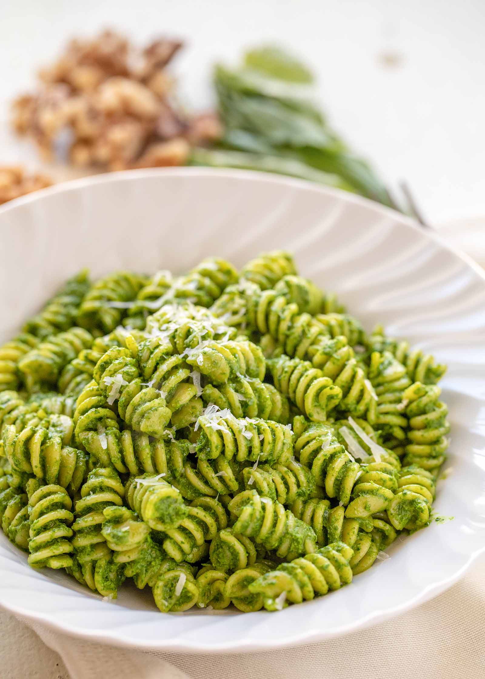 A white bowl with rotini and kale pesto inside. Walnuts and basil are in the background.
