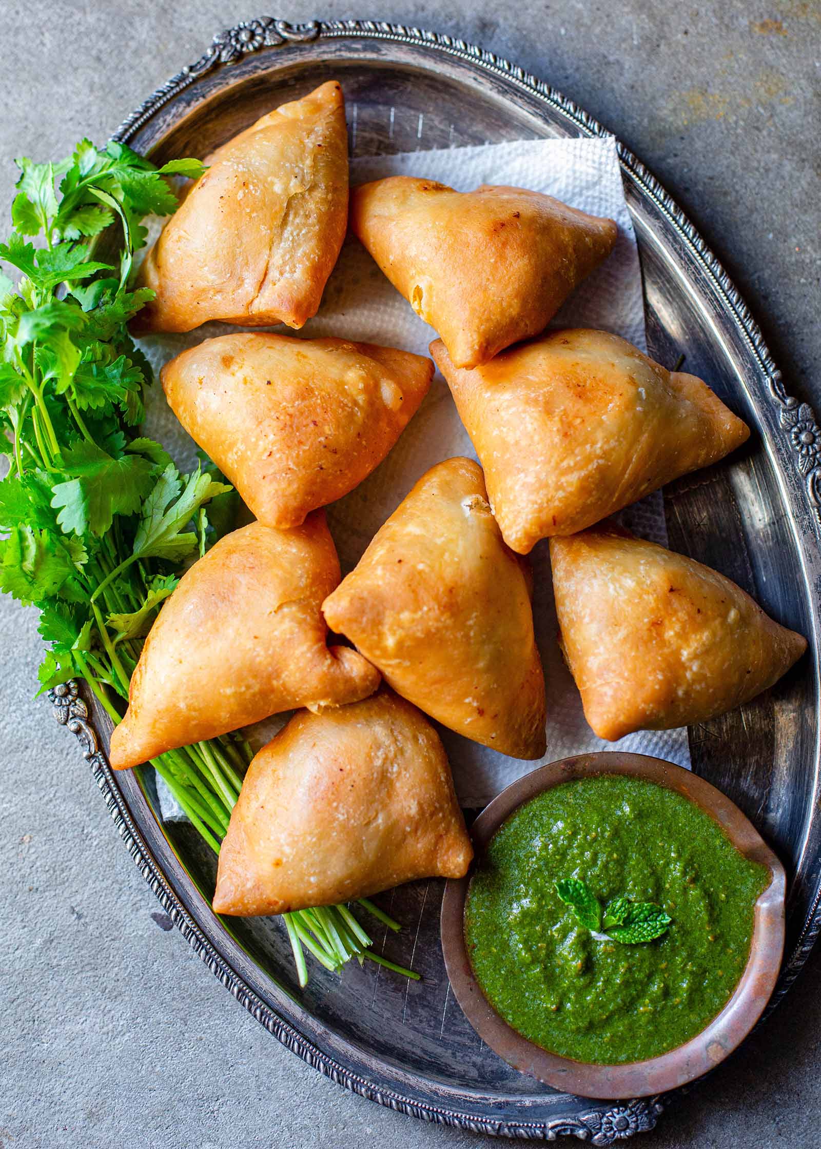 Vegetarian Samosas on a bed of herbs with one samosa filled with potato and peas alongside a cilantro mint chutney or dipping sauce in a small wooden bowl..
