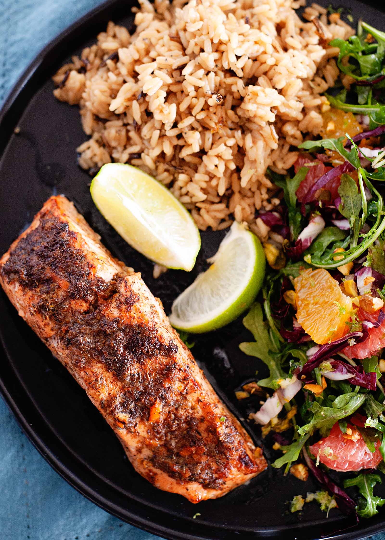 A black plate has a blackened fillet of jerk salmon on the lower left corner. Two lime wedges, red rice and a mixed green salad with citrus is also on the plate. The plate is on a blue linen.