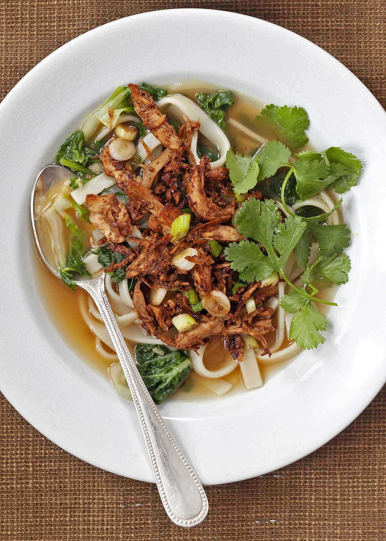Gochujang Chicken and Udon Noodle Soup in a white bowl with a spoon and fresh herbs.