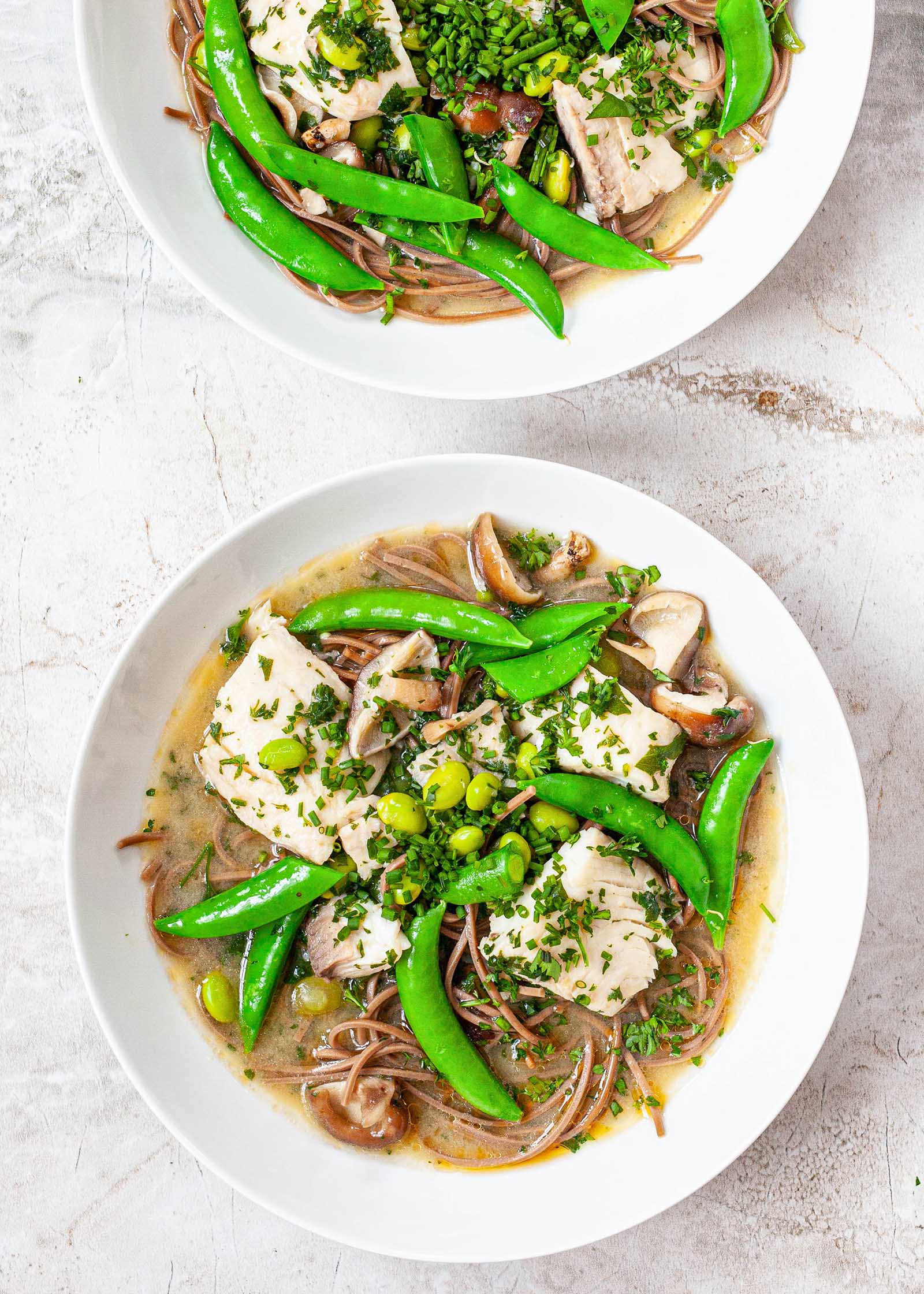 Two white bowls filled with quick and easy Miso Soup with fish, peas, herbs and soba noodles on a marble background.