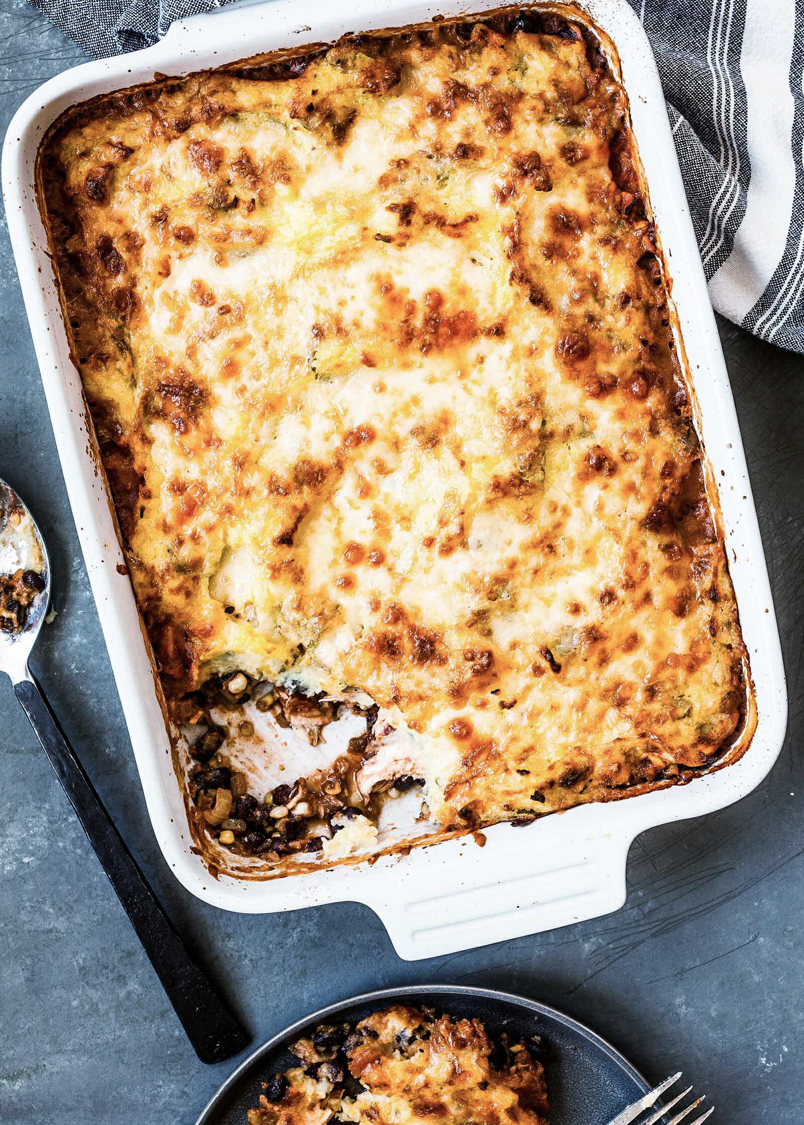 Weeknight shepherd's pie set vertically with a scoop missing and on plate below. Serving spoon and striped napkin set on either side of the dish.