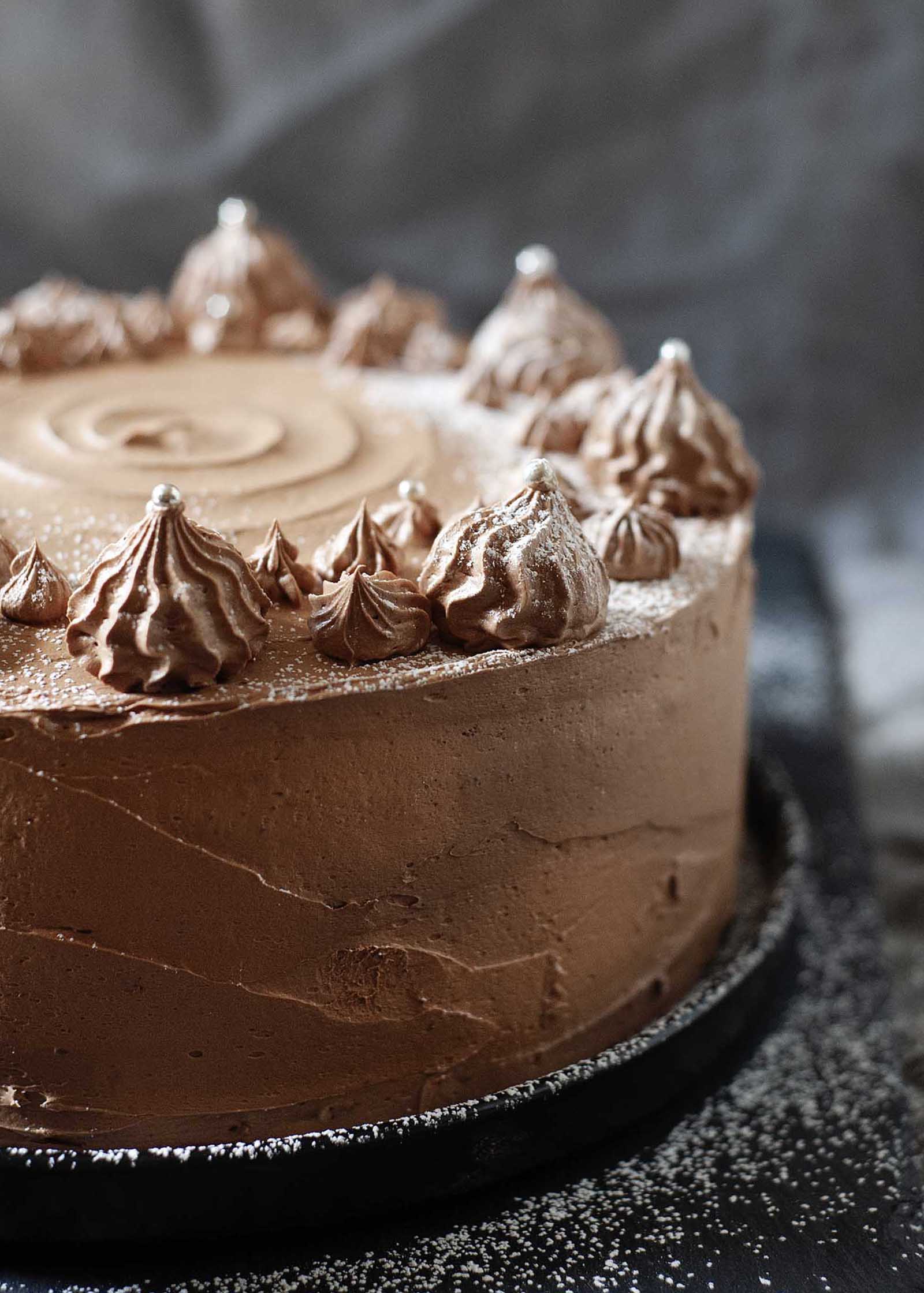 Whole choclate layer cake with chocolate swiss meringue buttercream swirls on top, dusted with powdered sugar against a dark background