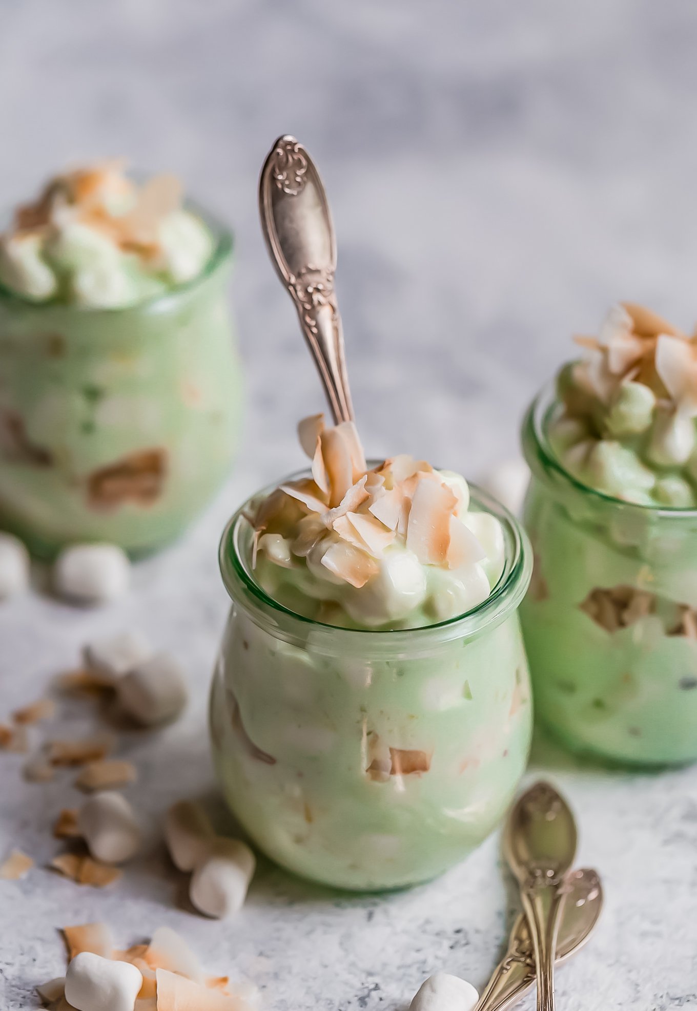 ¡La ensalada Watergate es el acompañamiento perfecto de Pascua! También conocida como Pistachio Fluff, esta receta de ensalada Watergate es esponjosa y deliciosa. ¡Toma una cuchara porque esta receta clásica de postres tiene un lugar en cada mesa, especialmente en las mesas festivas! tarros de ensalada watergate cubiertos con malvaviscos y coco tostado