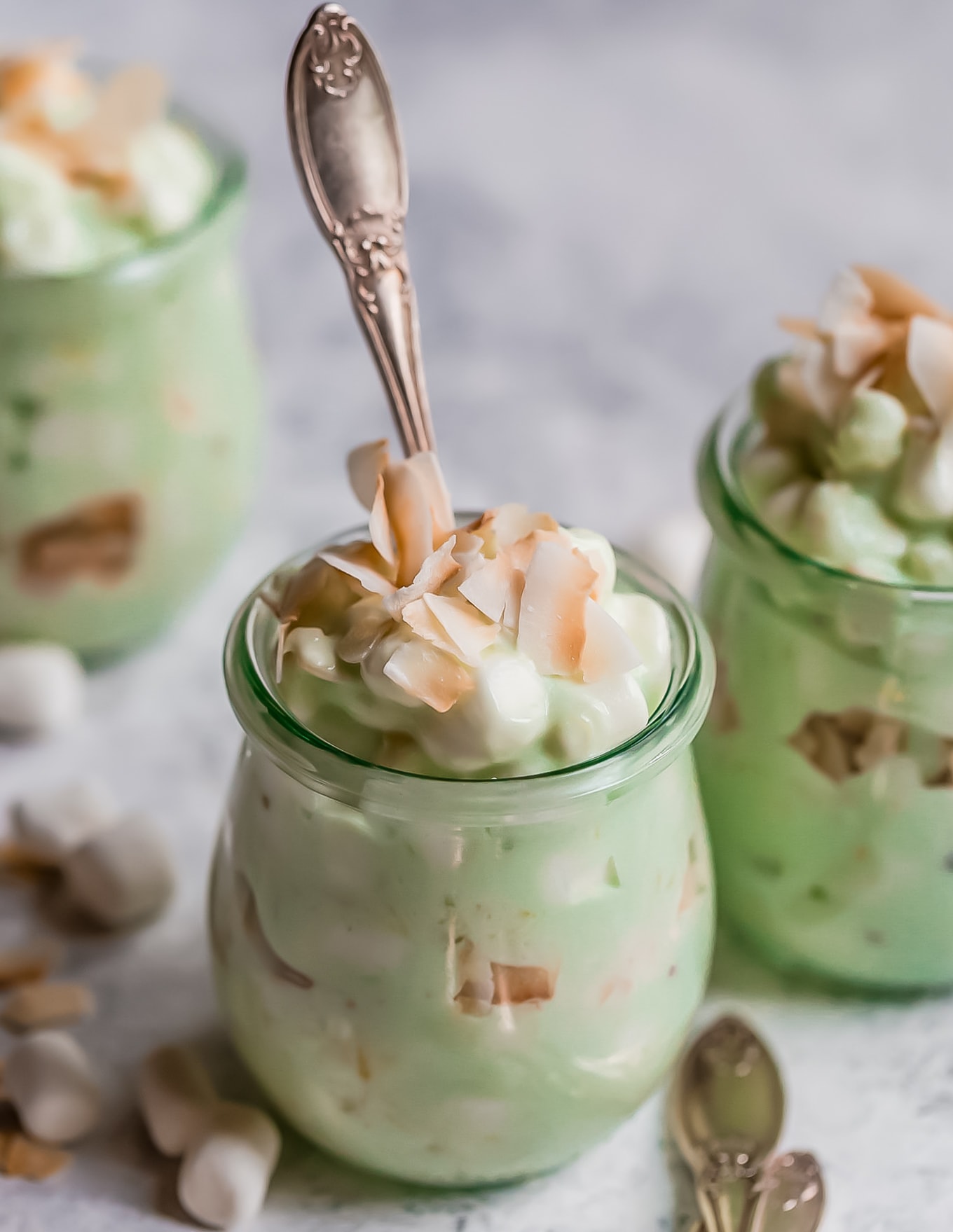¡La ensalada Watergate es el acompañamiento perfecto de Pascua! También conocida como Pistachio Fluff, esta receta de ensalada Watergate es esponjosa y deliciosa. ¡Toma una cuchara porque esta receta clásica de postres tiene un lugar en cada mesa, especialmente en las mesas festivas! pequeños frascos de ensalada watergate con una cuchara