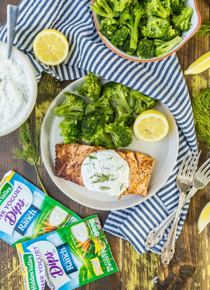 paesaggio con un piatto di salmone e broccoli, un piatto di broccoli, un canovaccio a strisce e pacchetti di ranch di yogurt greco