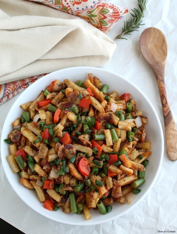 Grande insalata di patate bianche con patate fritte al rosmarino - L'insalata ha pezzi di patatine fritte, fagiolini a fette, peperone rosso, pezzi di pancetta e cipolle verdi tritate. la ciotola è su uno sfondo bianco con una biancheria da letto verde, blu e arancione, c'è un colino bianco con fagiolini freschi dietro e un cucchiaio da portata in legno