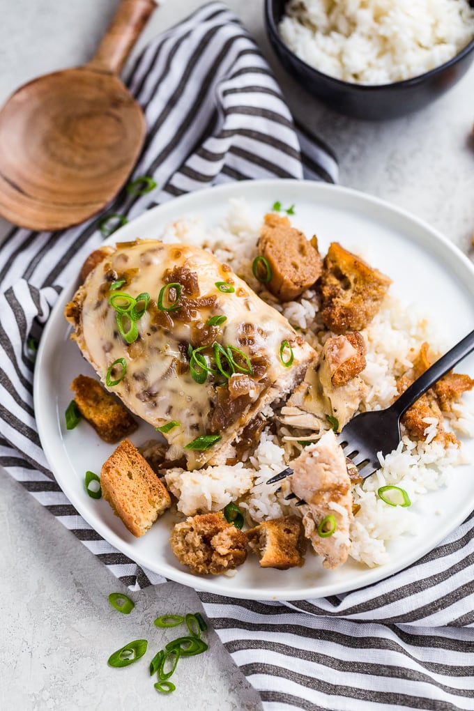 French Onion Chicken es una comida fácil de comer que encantará a toda la familia. Esta receta de pollo sartén sabe igual que su sopa favorita en forma de plato principal. Sopa de cebolla francesa El pollo es una pechuga de pollo con queso suizo, sopa de cebolla francesa, crotones y más queso. Pechuga de pollo cubierta de queso, en un plato blanco con arroz blanco y picatostes