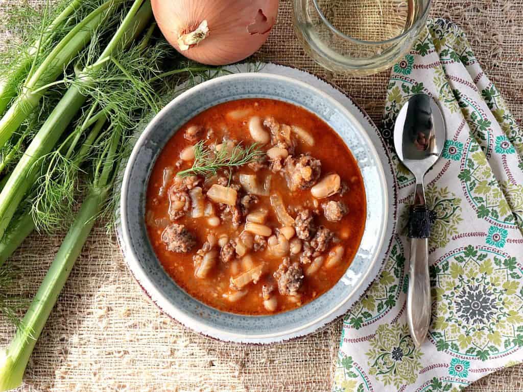 Questa deliziosa zuppa di fagioli bianchi, finocchio e salsiccia italiana farà sicuramente piacere a tutta la famiglia. La profondità del sapore della miscela di finocchio dolce e fagioli bianchi è un sontuoso trattamento per le papille gustative. - Renee's Kudos Kitchen Una ciotola di fagioli bianchi, finocchio e zuppa di salsiccia italiana con finocchio, cipolla e un bicchiere di vino. - www.kudoskitchenbyrenee.com