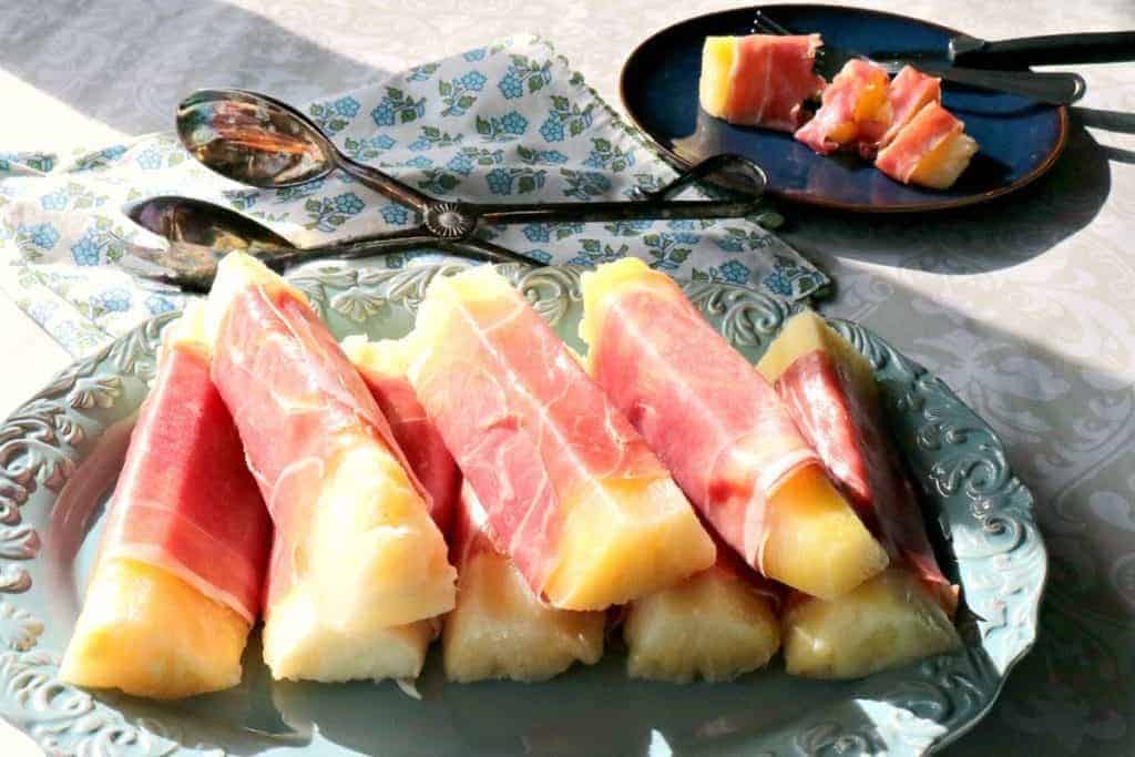 Riepilogo delle ricette del brunch Ananas avvolto con prosciutto su un piatto ovale blu con tenaglie in background.