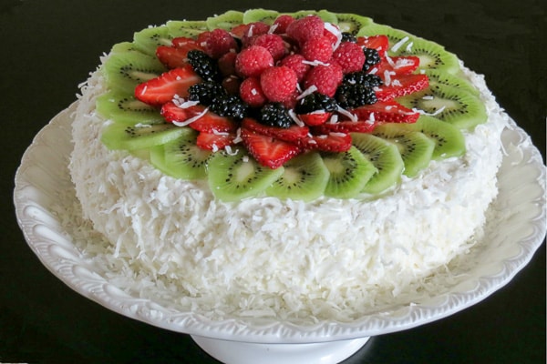 Una ricetta di pan di spagna soffice e ariosa fatta con strati di crema dolce e condita con cocco e frutta. Strati di pan di spagna con frutta su un piatto torta ricoperta di frutta.