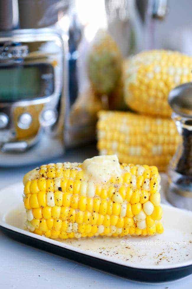 Corn on the cob with butter on top, on a dish with a slow cooker and two ears of corn in the background
