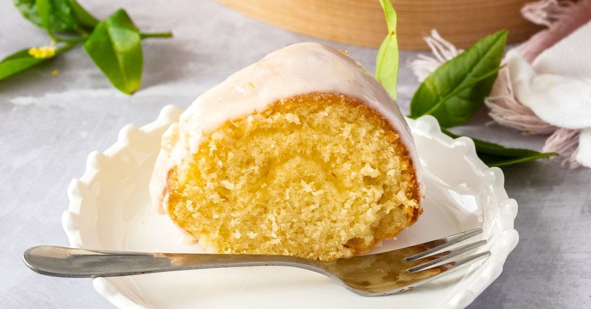 Uma fatia de bolo de manteiga de baunilha em um prato de bolo branco com um garfo na frente. Algumas folhas de limão ficam ao seu redor