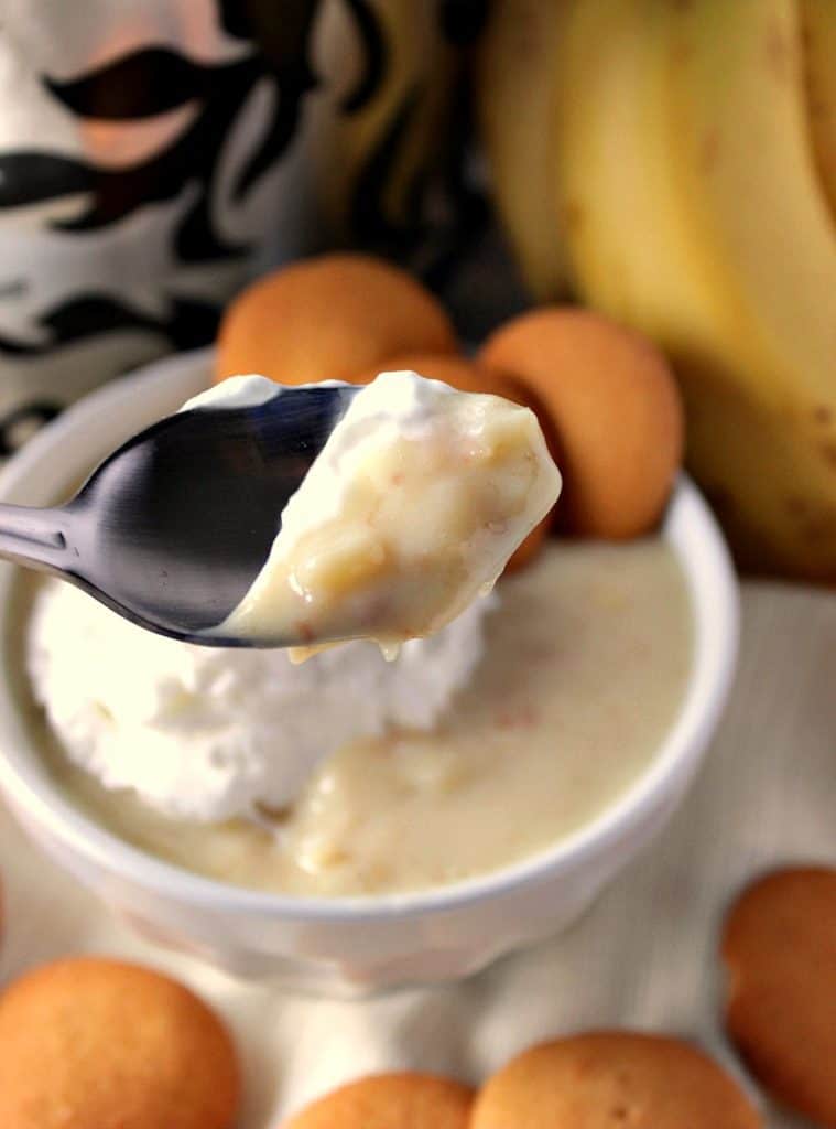 Close-up de pudim de banana em uma colher com uma tigela por baixo, além de pudim, chantilly e bolacha de baunilha.