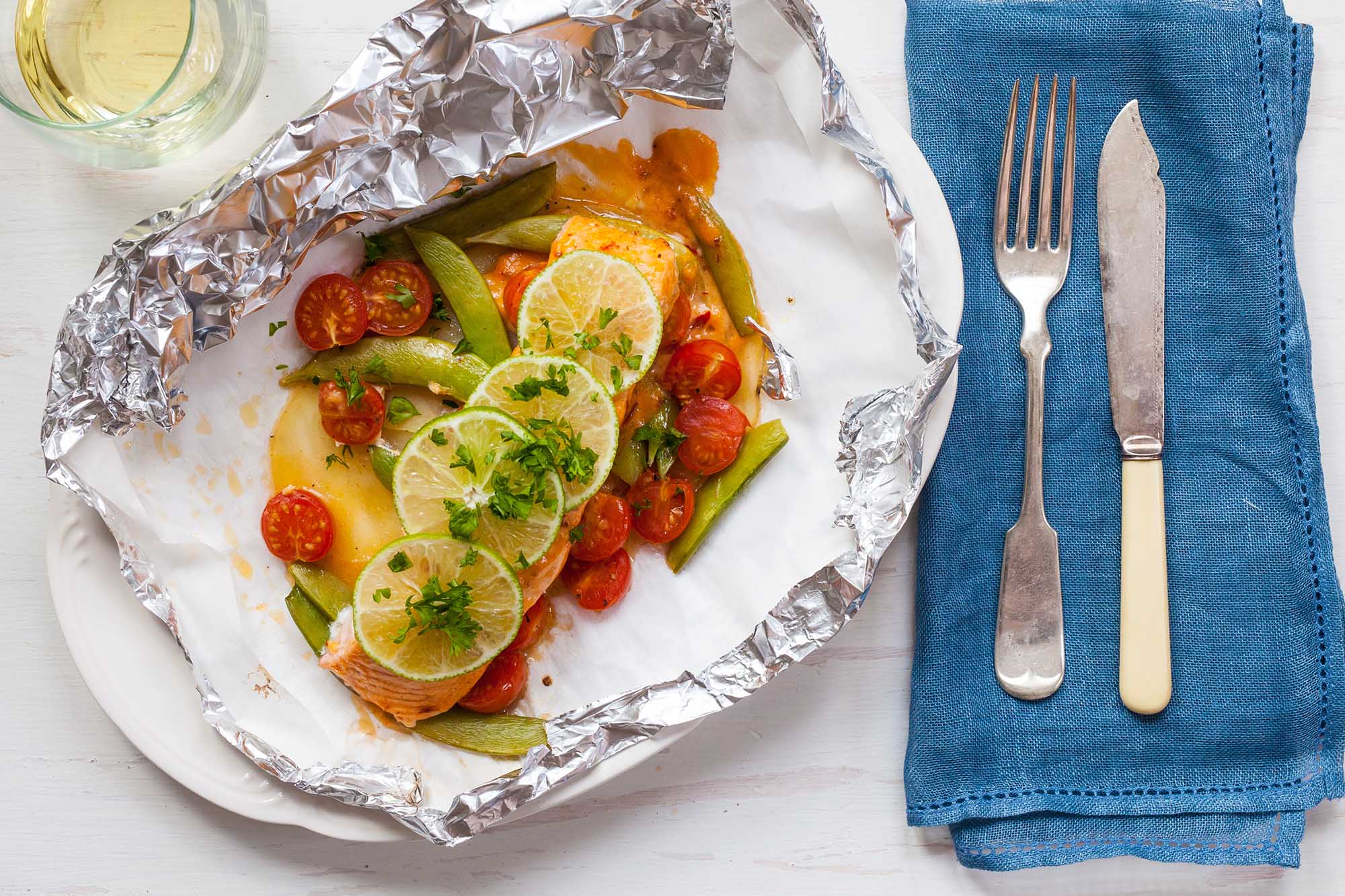 Um prato contém um pacote de papel alumínio para revelar salmão saudável com tomates, ervilhas e limão fatiado por cima. Um guardanapo azul está à direita do pacote de papel alumínio e um garfo e faca estão no npakin.