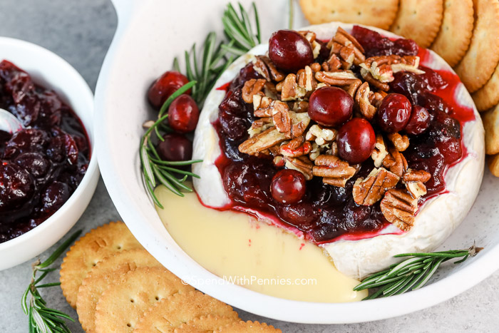 Brie assado coberto com molho de cranberry-noz, com queijo escorrendo e bolachas ao lado. 