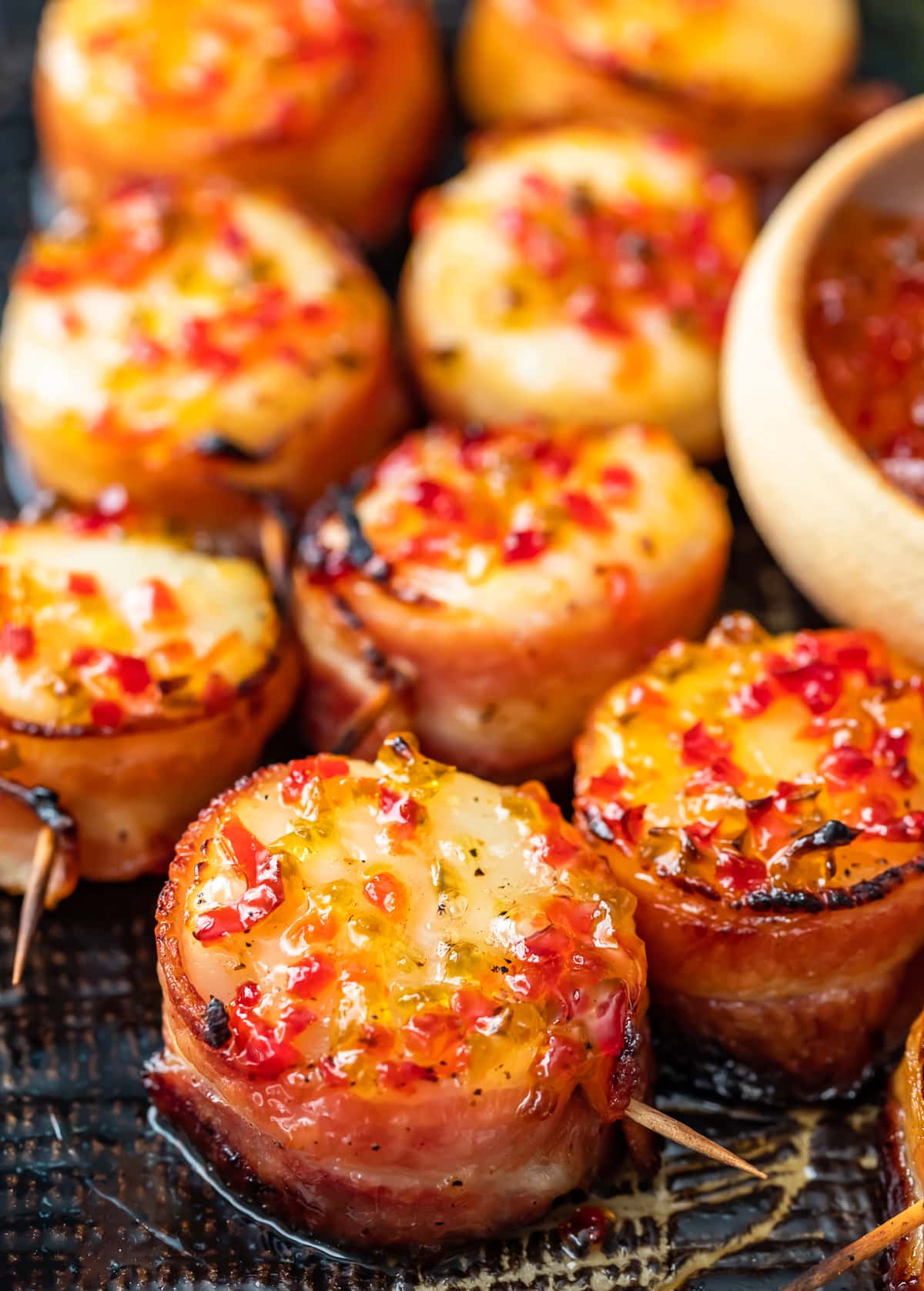 Las vieiras envueltas en tocino son un delicioso aperitivo para cualquier fiesta u ocasión especial. Tienen un sabor salado y mantecoso, con un poco de especias gracias a la gelatina de pimiento picante en la parte superior. ¡Esta receta de vieiras envueltas en tocino horneado es tan fácil y deliciosa! vieiras envueltas en tocino y cubiertas con gelatina de pimiento picante