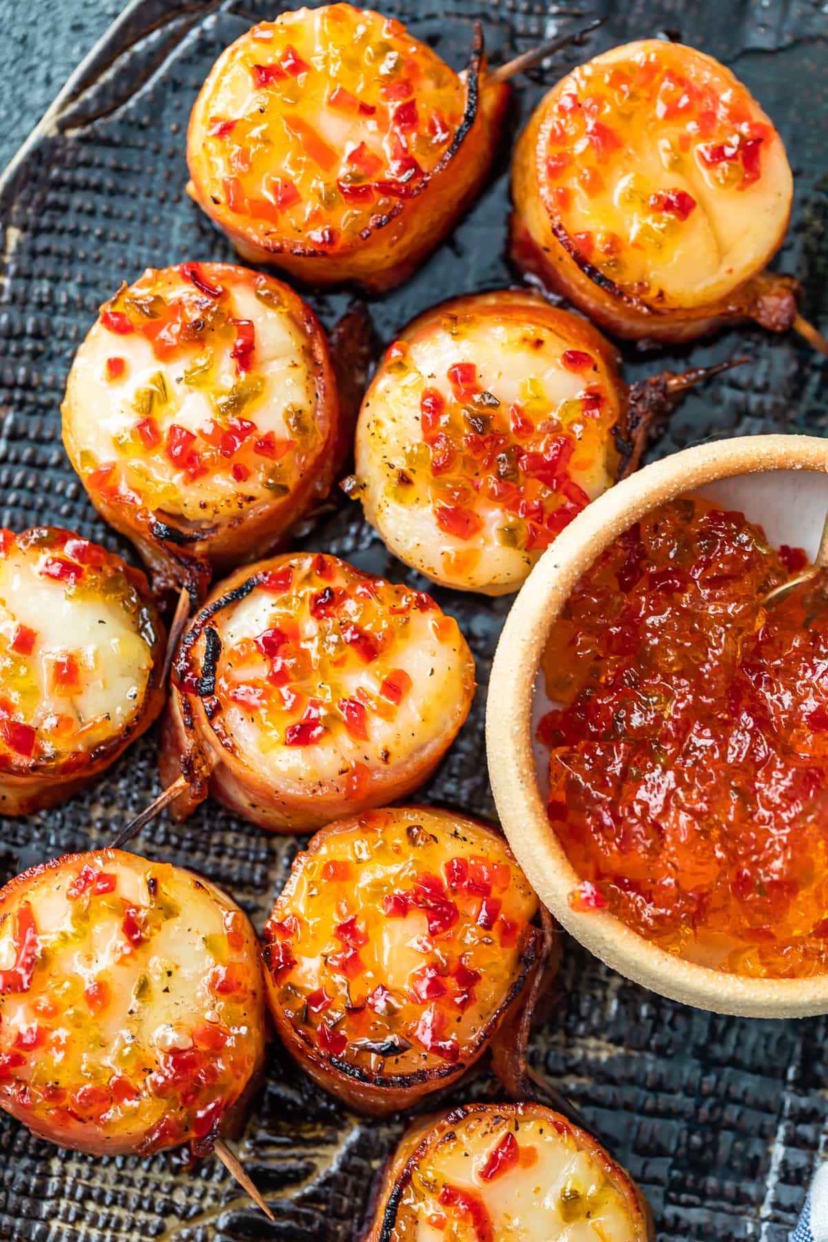 Las vieiras envueltas en tocino son un delicioso aperitivo para cualquier fiesta u ocasión especial. Tienen un sabor salado y mantecoso, con un poco de especias gracias a la gelatina de pimiento picante en la parte superior. ¡Esta receta de vieiras envueltas en tocino horneado es tan fácil y deliciosa! Vista aérea de un plato de vieiras envueltas en tocino