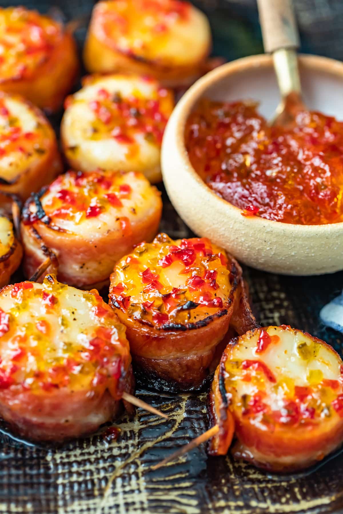 Las vieiras envueltas en tocino son un delicioso aperitivo para cualquier fiesta u ocasión especial. Tienen un sabor salado y mantecoso, con un poco de especias gracias a la gelatina de pimiento picante en la parte superior. ¡Esta receta de vieiras envueltas en tocino horneado es tan fácil y deliciosa! vieiras envueltas en tocino dulce y picante