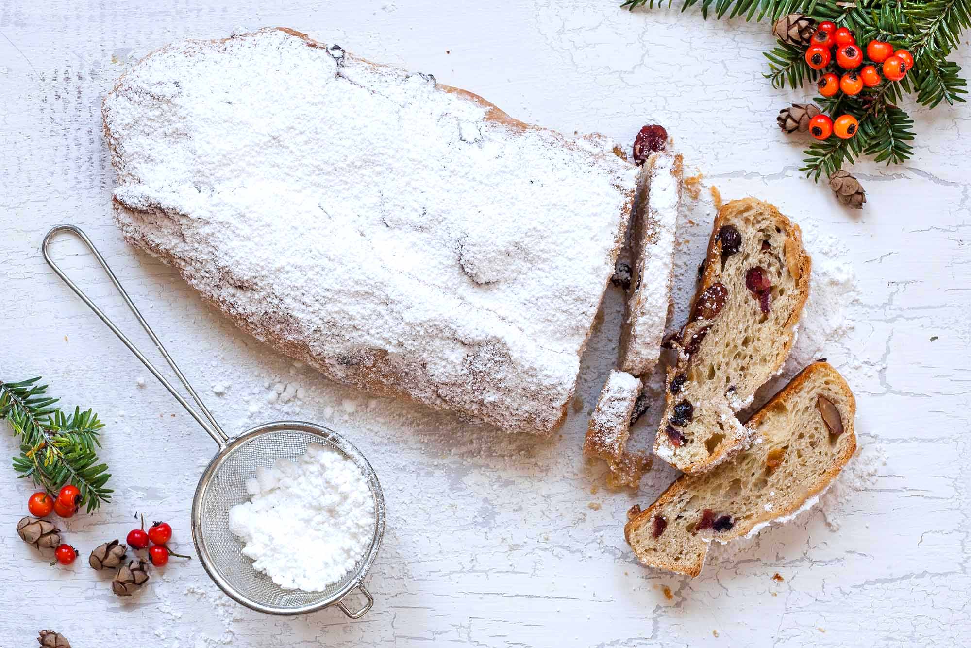 Stollen Christmas Pan cheia de frutas secas embebidas em rum revestidas com açúcar em pó e cortadas em uma bandeja com uma peneira de malha fina com açúcar em pó nas proximidades.