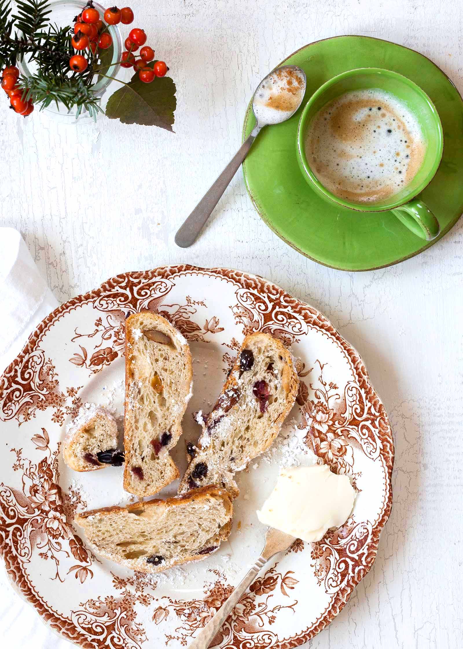 Pão de Natal Stollen recheado com frutas secas embebidas em rum, coberto de açúcar em pó e fatiado em um prato com uma peneira de malha fina com açúcar em pó e uma xícara de café em uma caneca verde.