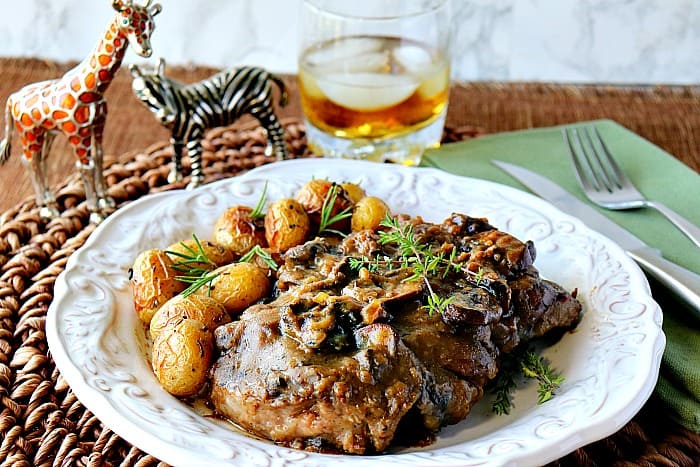 Bife com molho de cogumelos em um prato branco com batatas bebê e um copo de bourbon nas rochas no fundo.