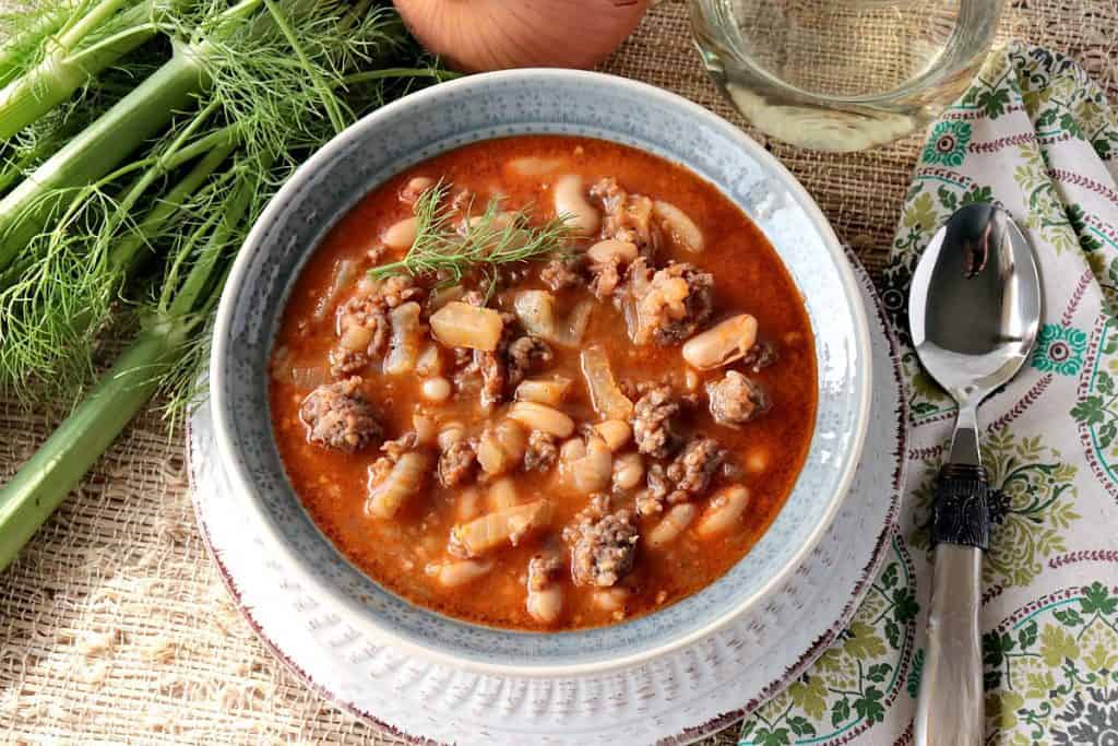 Uma tigela de feijão branco, erva-doce e sopa de linguiça italiana com erva-doce, cebola e um copo de vinho. 