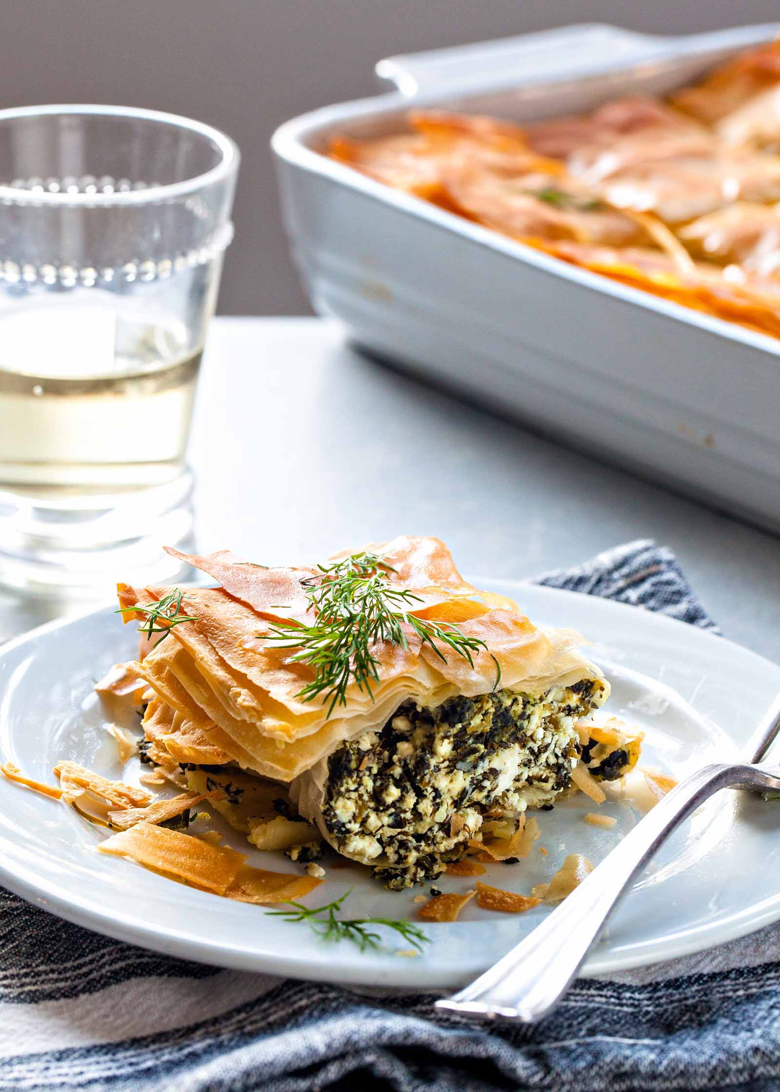 Fatia de torta de espinafre grego em um prato branco, com um garfo, um copo de vinho branco e a caçarola ao fundo,