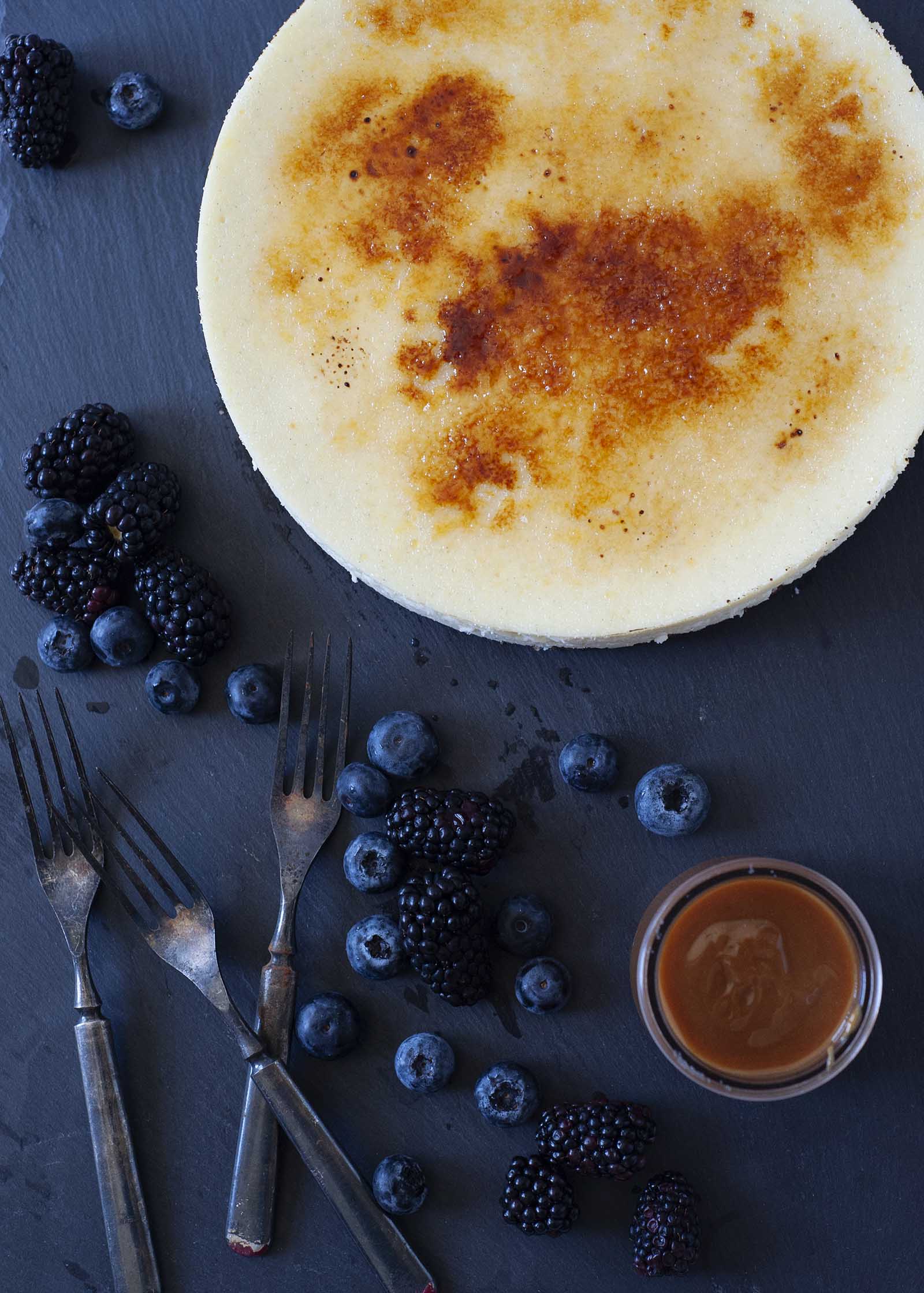 Cheesecake de baunilha com cobertura de açĂșcar crocante em uma lousa com frutas e caramelo nas proximidades.