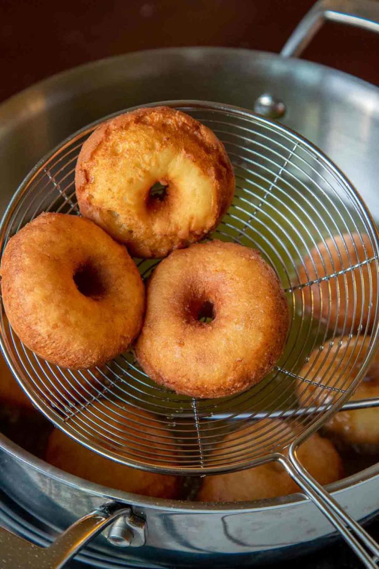 Uma deliciosa receita caseira de mini rosquinha lisa e arejada, com a melhor massa de rosquinha com queijo ricota e sem fermento necessário. Os donuts estão completamente prontos em apenas 30 minutos. Três donuts fritos em uma peneira ao lado de uma panela com donuts fritos.