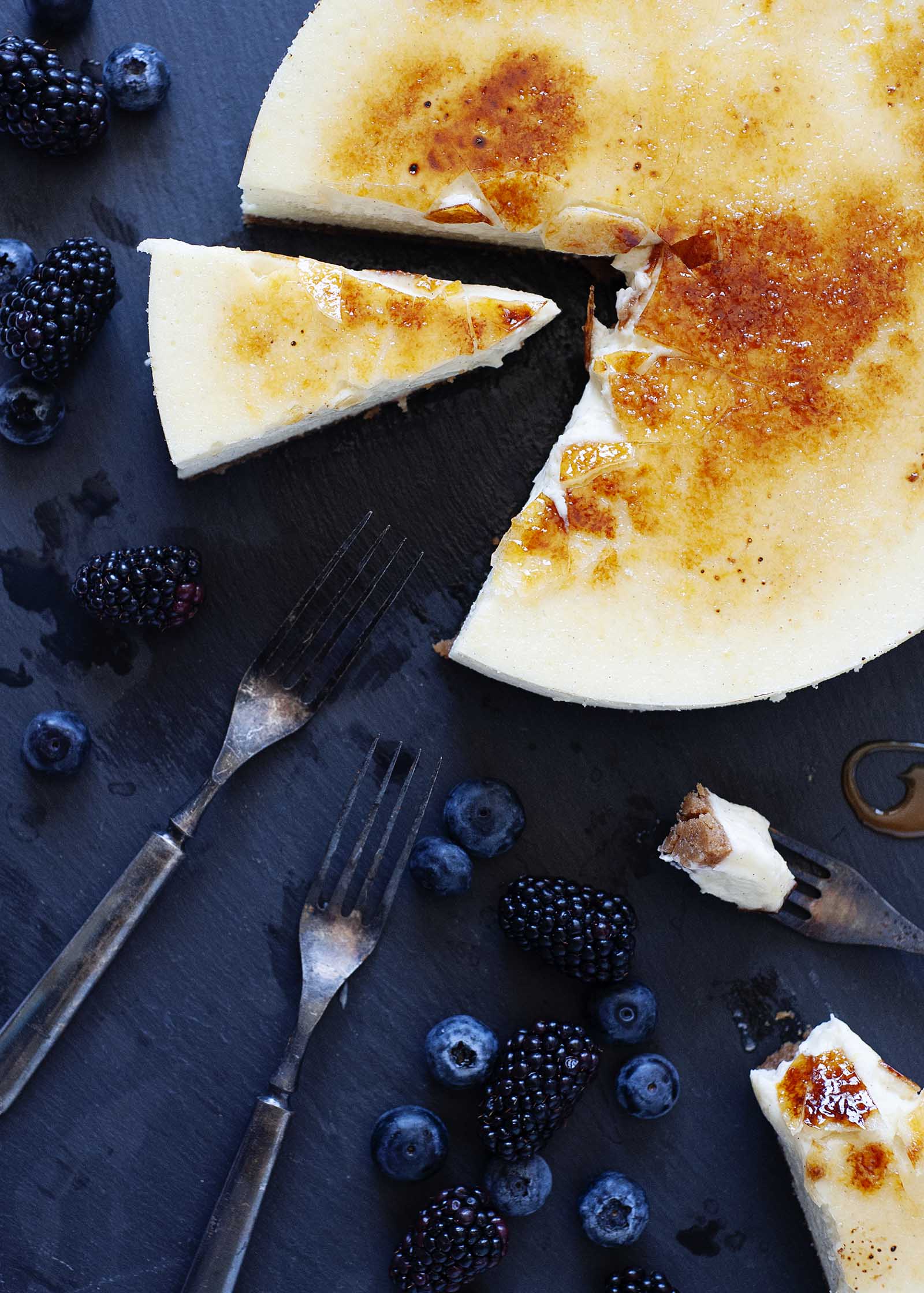 Cheesecake de baunilha com cobertura de açĂșcar frĂĄgil em uma placa de ardĂłsia com uma fatia cortada e frutas e garfos prĂłximos.